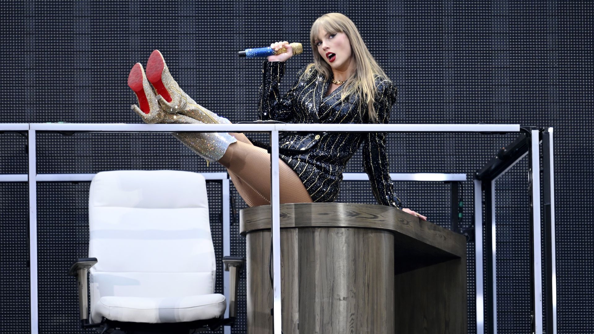 Taylor Swift op het podium in het Wembley Stadium op 21 juni 2024. Ze draagt sprankelende gouden laarzen en een met juwelen bezette blazer en zit op een bureau