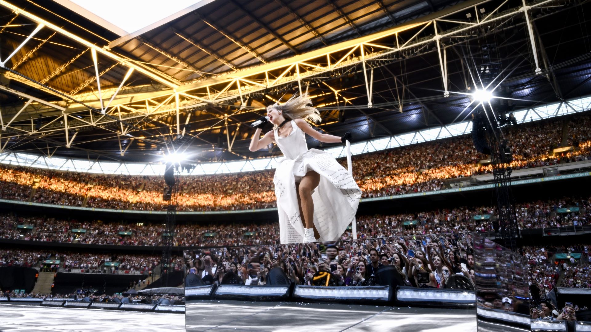 Taylor Swift op het podium in het Wembley Stadium op 23 juni 2024. Ze draagt een jurk bedekt met handgeschreven zwarte tekst en staat op een spiegelende platform