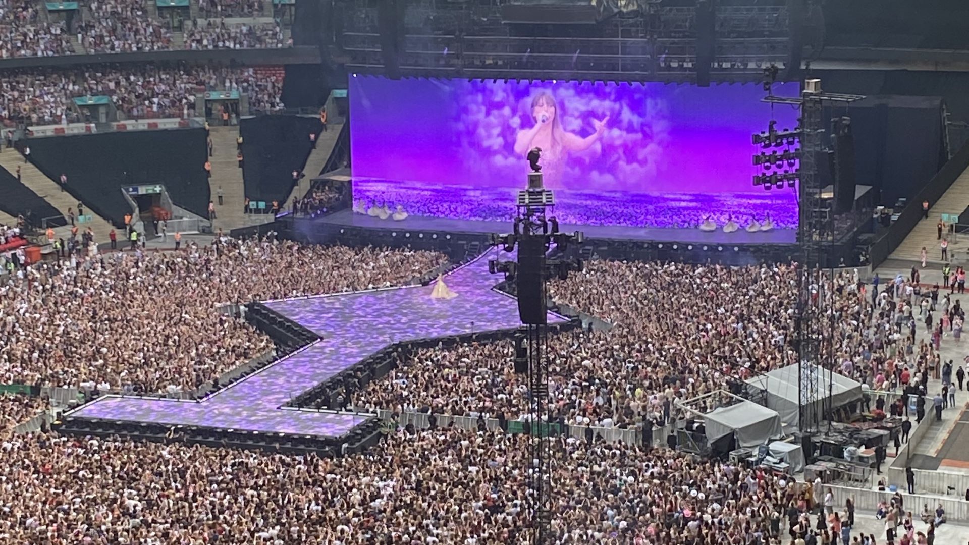 Een afbeelding van The Eras Tour, Londen, avond 1. Het stadion is vol en Taylor Swift is te zien op een groot scherm achter een loopbrug. Het uitzicht is enigszins geblokkeerd door een speaker